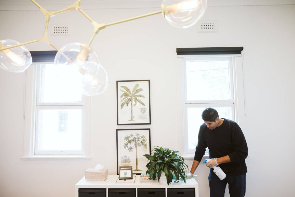 Man cleaning and dusting framed art and plants on a white shelf in a bright, minimalist interior space with large windows.